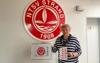 Bettina Ossyra, Geschäftsstellenleiterin beim NTSV Strand 08 präsentiert die eigene Briefmarke mit dem Vereinslogo des Timmendorfer Sportclubs.
